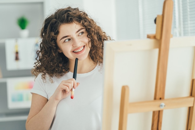 A mulher atraente com um pincel de arte pintando um quadro no cavalete