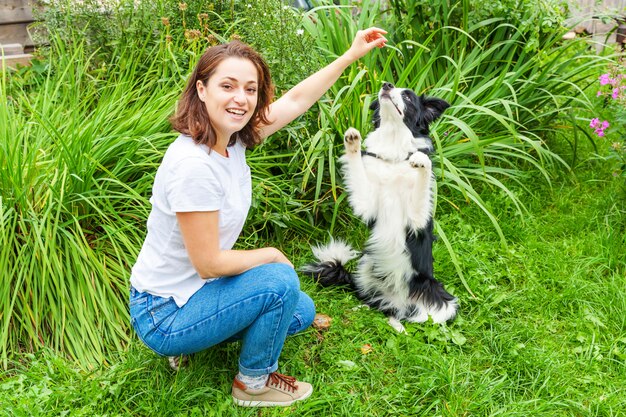 A mulher atrativa nova de sorriso que joga com cão de cachorrinho bonito border collie no jardim do verão ou a cidade estacionam o fundo exterior. Truque de treinamento de garota com amigo de cachorro. Conceito de cuidados e animais de estimação.