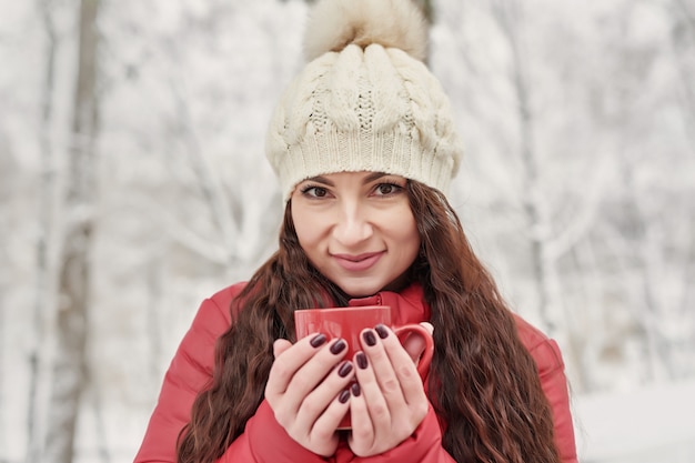 A mulher bebe o chá ou o café quente da xícara no jardim acolhedor da casa nevado na manhã do inverno. Mulher bonita, aproveitando o inverno ao ar livre com uma caneca de bebida quente. Férias de natal. Estilo de vida aconchegante no inverno.