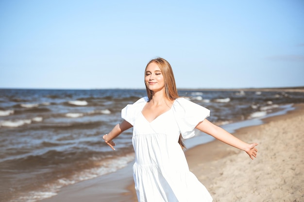 A mulher bonita de sorriso feliz está na praia do oceano em um vestido branco do verão, braços abertos.