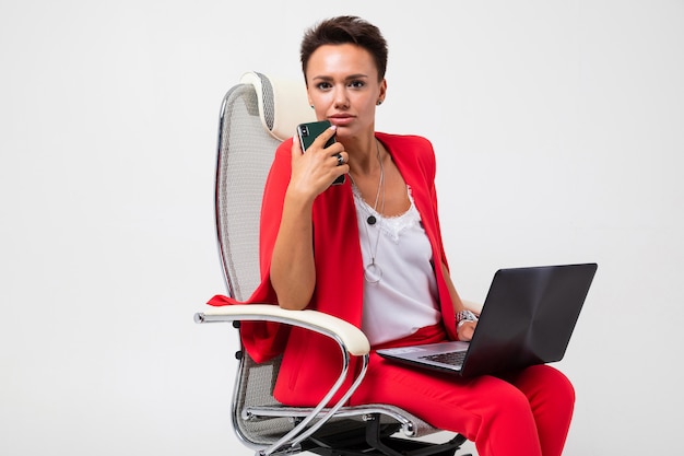 A mulher bonita no terno vermelho do escritório com cabelo escuro curto faz uma reunião importante e trabalha com o laptop, senta-se em uma cadeira