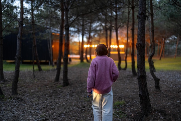 A mulher caminha na floresta de pinheiros perto da cabana de madeira ao entardecer