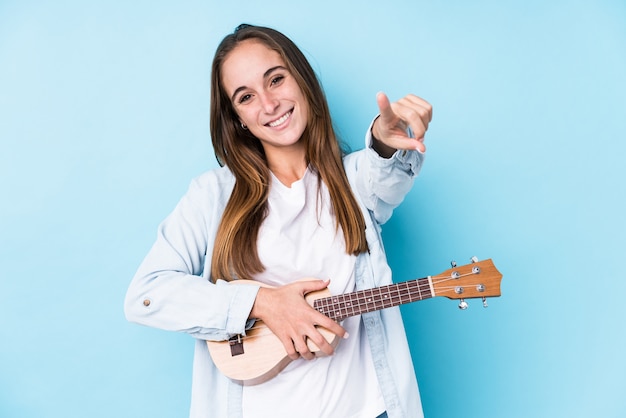 A mulher caucasiano nova que guarda um ukelele isolou os sorrisos alegres que apontam para a parte dianteira.