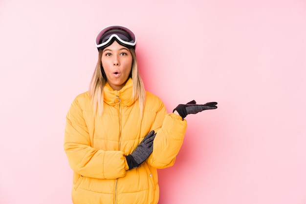 A mulher caucasiano nova que veste um esqui veste-se em uma parede cor-de-rosa imprimiu guardar o espaço da cópia na palma.