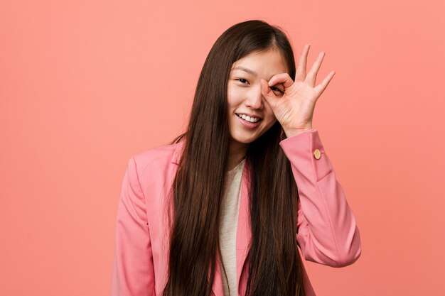 A mulher chinesa do negócio novo que veste o terno cor-de-rosa excitou mantendo o gesto aprovado no olho.