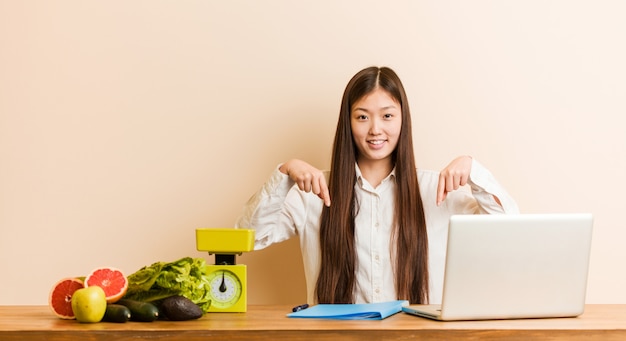 A mulher chinesa do nutricionista novo que trabalha com seu portátil aponta para baixo com os dedos, sentimento positivo.