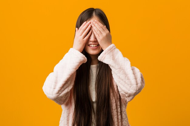 A mulher chinesa nova no pijama cobre os olhos com as mãos, sorrisos amplamente à espera de uma surpresa.