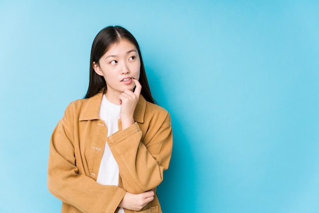 A mulher chinesa nova que levanta em uma parede azul relaxou o pensamento sobre algo que olha um espaço em branco.