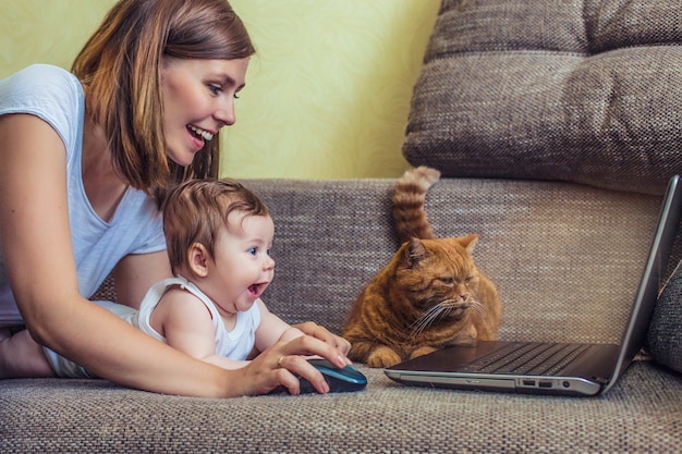 A mulher com um bebê e um gato no laptop deitada no sofá