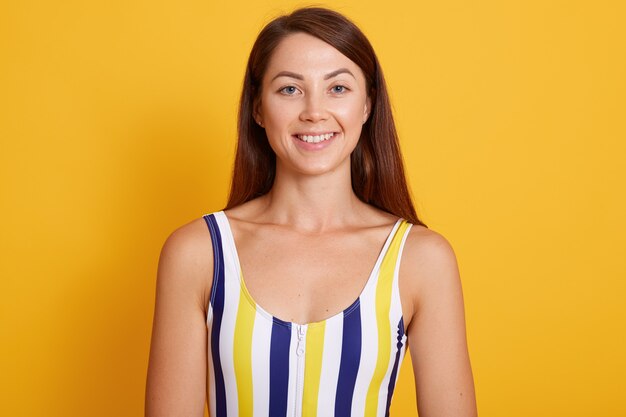 A mulher de cabelo escura bonita vestida o terno de natação listrado está sorrindo. levantamento fêmea isolado sobre a parede amarela. conceito de pessoas.