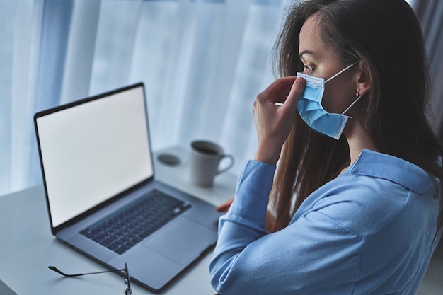 A mulher de negócios usa uma máscara de proteção médica on-line trabalhando em casa, no laptop durante o auto-isolamento e quarentena.