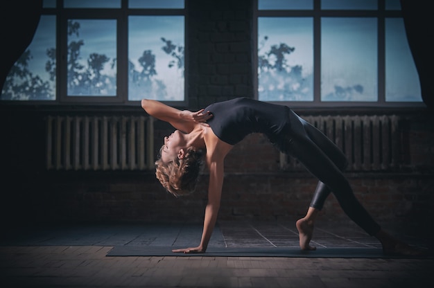 A mulher desportiva bonita do ajuste yogini pratica o asana da ioga Pose da coisa selvagem no salão escuro