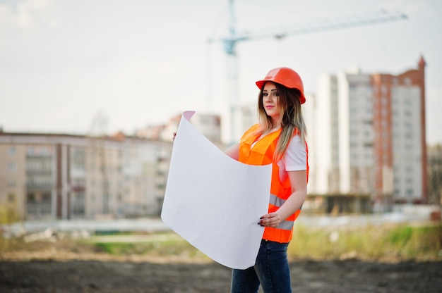 A mulher do construtor do coordenador no colete uniforme e no capacete protetor alaranjado mantem o papel de negócio contra construções novas com guindaste.