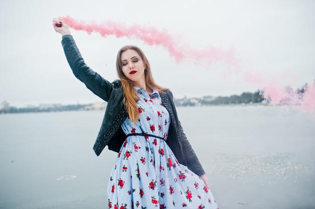 A mulher elegante na jaqueta de couro mantém o reflexo de fumaça rosa em dia de inverno contra lago congelado.