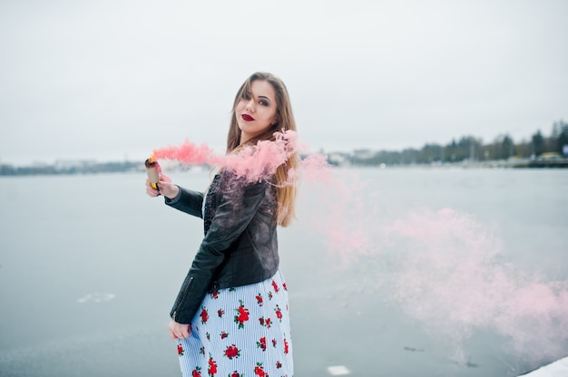 A mulher elegante na jaqueta de couro mantém o reflexo de fumaça rosa em dia de inverno contra lago congelado.