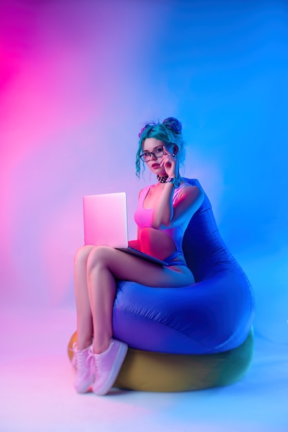 A mulher em um maiô brilhante com cabelo azul está sentada em uma bolsa de cadeira com um laptop