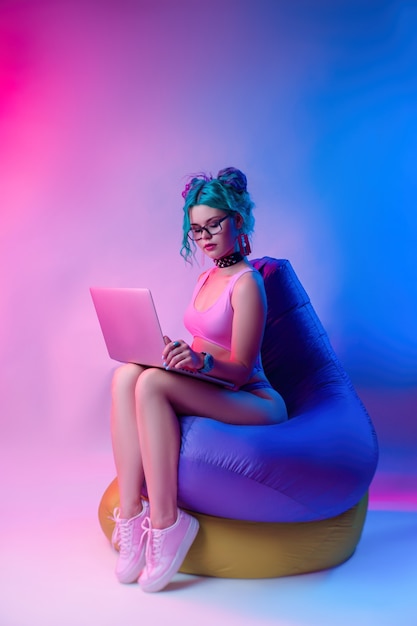 A mulher em um maiô brilhante com cabelo azul está sentada em uma bolsa de cadeira com um laptop