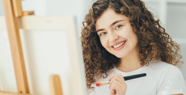 Foto a mulher encaracolada com um pincel de arte pintando um quadro no cavalete