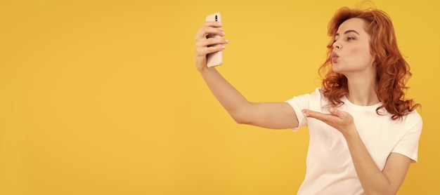 A mulher faz um gesto de beijo de mão para a câmera do telefone móvel fundo amarelo selfie Mulher isolada banner de retrato de rosto com espaço de cópia simulado
