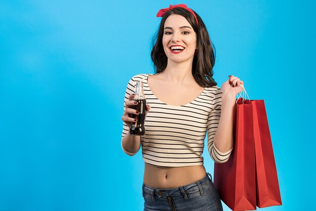 A mulher feliz com sacos bebendo uma coca-cola no fundo azul