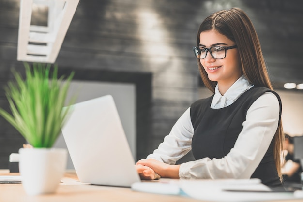 A mulher feliz com um laptop moderno sentada na frente da mesa