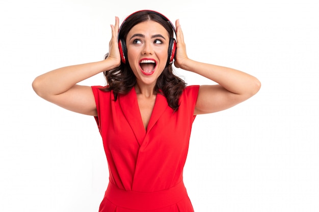 A mulher feliz sorri amplamente em uma superfície branca, senhora elegante em um vestido vermelho ouve música com fones de ouvido