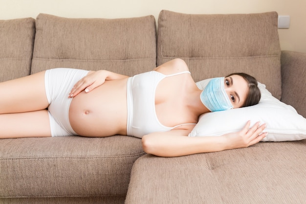 A mulher grávida está usando máscara médica protetora para se proteger e o futuro bebê contra o coronavírus. mãe está deitada no sofá em casa. conceito de quarentena global