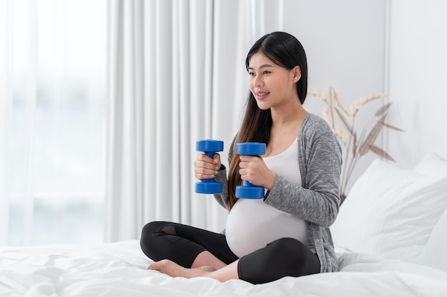 A mulher grávida feliz asiática está sentada e se exercita na cama, gravidez, maternidade, pessoas e conceito de expectativa