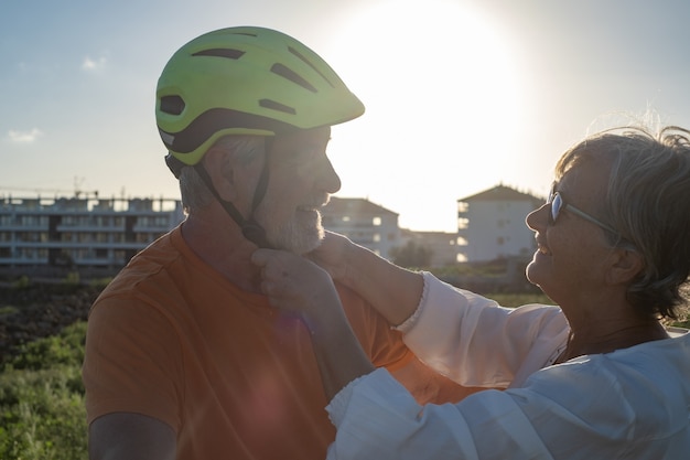 A mulher idosa cuida do marido ciclista colocando seu capacete. Luz forte do pôr do sol