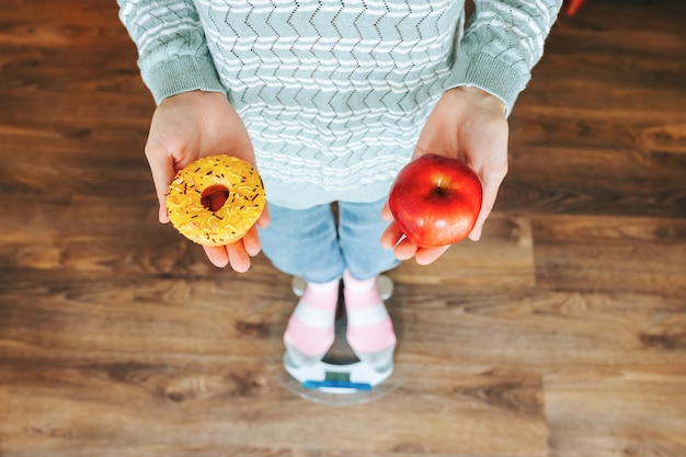 Foto a mulher irreconhecível fica na balança e faz uma escolha difícil de rosquinha ou excesso de peso de maçã d