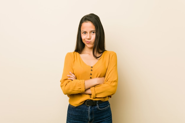 Foto a mulher latino-americano nova contra um fundo bege que olha carrancuda no descontentamento, mantém os braços dobrados.