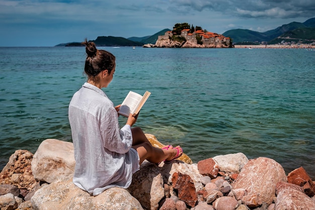 A mulher lê o livro na beira-mar em Sveti Stefan. Montenegro