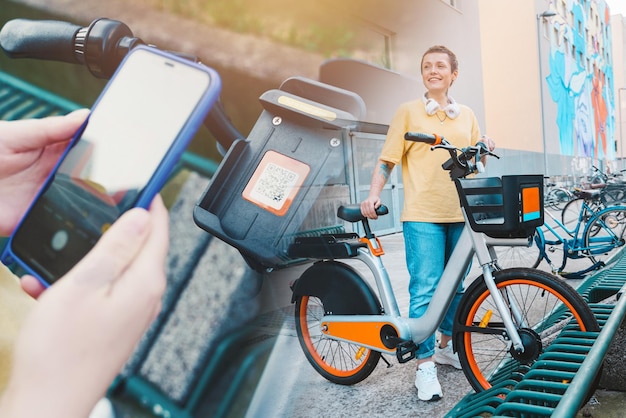 A mulher leva uma bicicleta alugada em um estacionamento de bicicletas com um smartphone