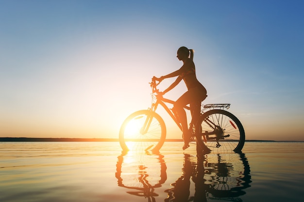 A mulher loira forte em um terno colorido senta-se na bicicleta na água ao pôr do sol em um dia quente de verão. Conceito de aptidão.