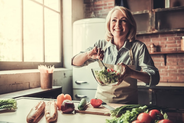 Foto a mulher madura bonita no avental está misturando a salada.