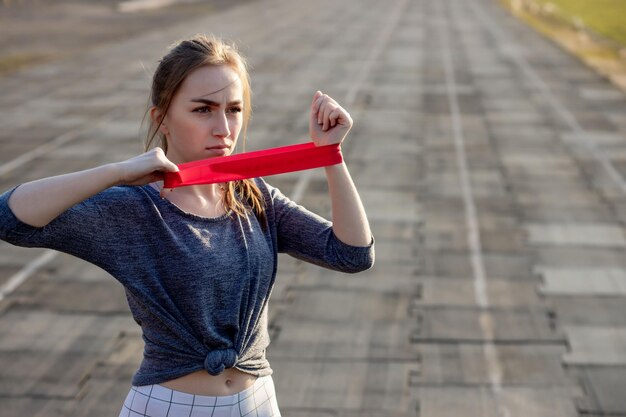 A mulher magro nova no sportswear que faz agachamentos exercita com elástico em uma trilha revestida preta do estádio