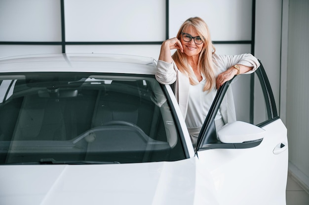 A mulher moderna do automóvel na roupa formal branca está na concessionária de automóveis