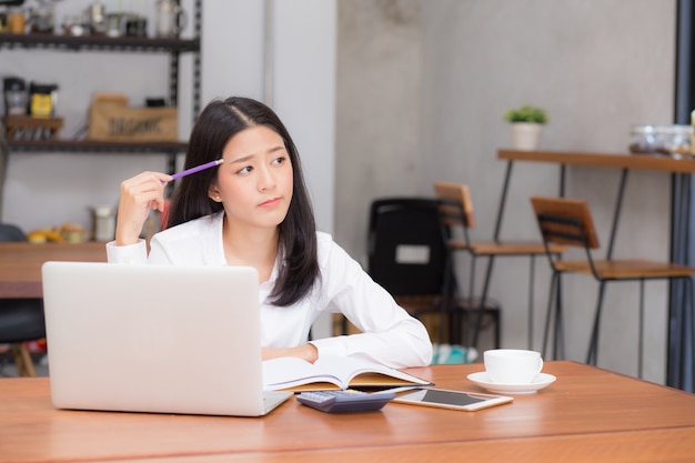 A mulher nova asiática bonita que trabalha em linha com portátil e pensa o projeto para a ideia na loja moderna do café.