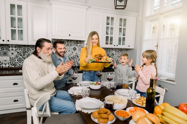 A mulher que guarda a bandeja com peru assado e decora para o jantar em família em casa. Todos os membros da família estão sentados à mesa, animados, felizes e aplaudem