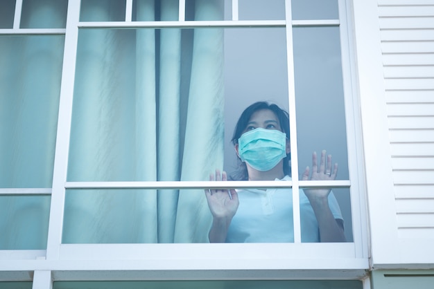 A mulher que usava uma máscara médica ficou olhando tristemente para a janela em casa, para quarentena
