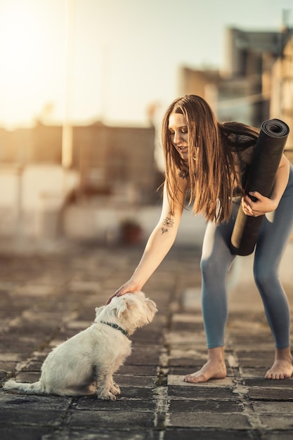 A mulher relaxada está enrolando a esteira de exercícios e se preparando para praticar ioga em um terraço na cobertura, apoiada por seu cachorro de estimação ao pôr do sol.