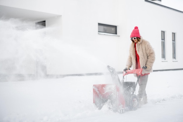 A mulher remove a neve com uma máquina do lançador de neve perto da casa