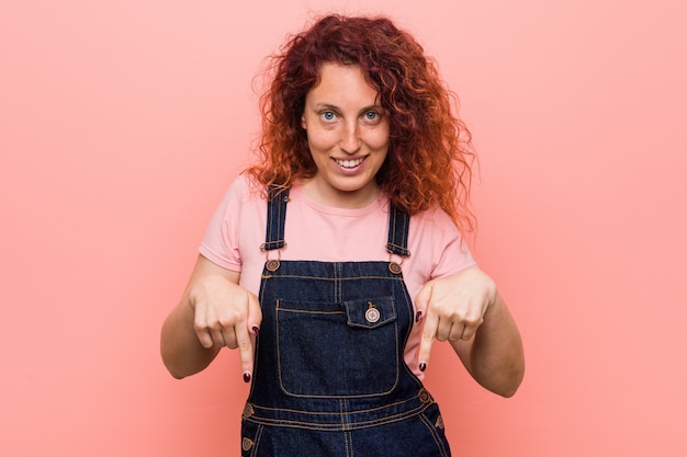 A mulher ruiva bonita nova do gengibre que veste um dungaree das calças de brim aponta para baixo com os dedos, sentimento positivo