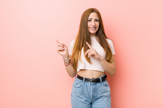 A mulher ruiva nova do gengibre contra uma parede cor-de-rosa chocou apontar com indicadores para um espaço da cópia.