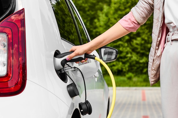 A mulher segura a fonte de alimentação conectada a um carro elétrico na estação de carregamento pública ao ar livre.