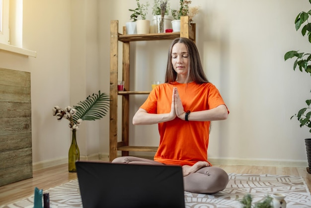 A mulher senta-se em posição de lótus no chão em frente a um laptop medita e relaxa enquanto ouve uma palestra online ou em vídeo Conceito de uma sessão de terapia remota meditação e ioga