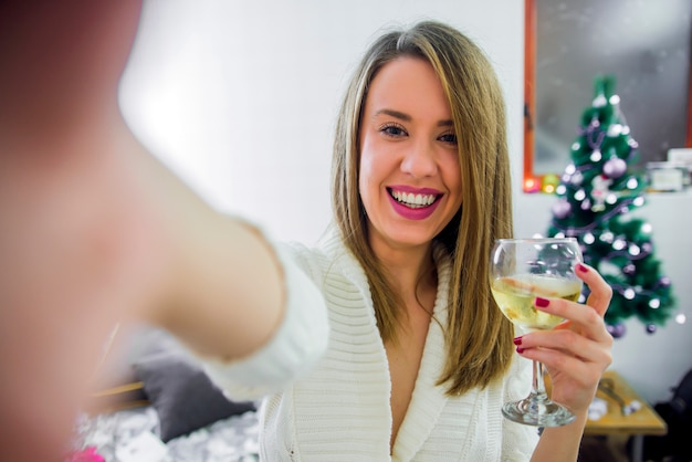 A mulher toma o selfie no telefone com a árvore de natal abrace o copo de videira, comemorando o ano novo. conceito de natal - mulher jovem que toma foto de selfie perto de árvore de natal decorada