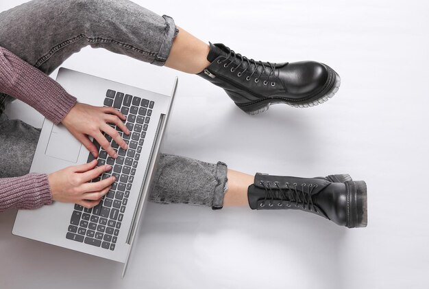 A mulher usa um laptop em um fundo branco pernas femininas em jeans e botas trabalho online vista superior