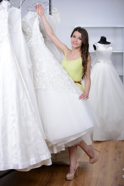 A noiva bonita de sorriso escolhe o vestido branco na loja do casamento.