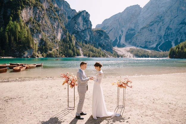 A noiva coloca um anel para o noivo no local da cerimônia, com um arco de colunas florais de outono, tendo como pano de fundo o lago di braies, na itália